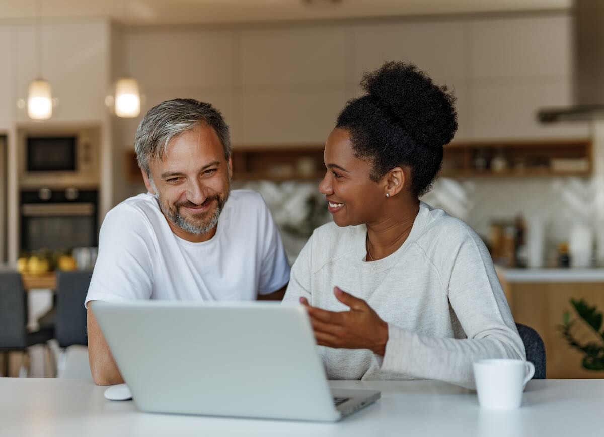 Diverse couple sitting at a kitchen looking at a laptop